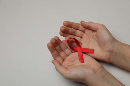 Little girl holding red ribbon on beige background, closeup with space for text. AIDS disease...