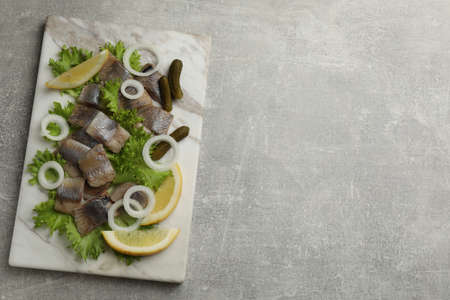 Sliced salted herring fillet served with lettuce, pickles, onion rings and lemon on gray table, top...
