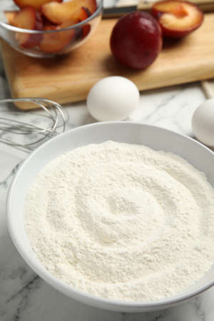 Bowl with flour on white marble table, closeup. Cooking of delicious plum cake
