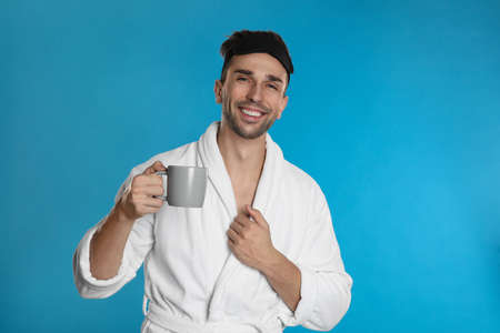 Young man in bathrobe with cup of coffee on light blue backgroundの写真素材