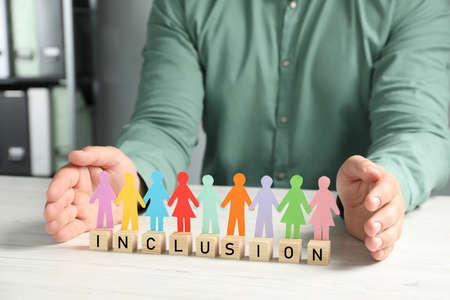 Man protecting paper human figures and wooden cubes with word Inclusion at table indoors, closeupの写真素材