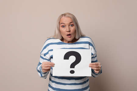 Emotional mature woman holding paper with question mark on beige backgroundの写真素材