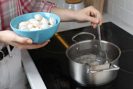 Woman putting frozen dumplings into saucepan with boiling water on cooktop in kitchen, closeup