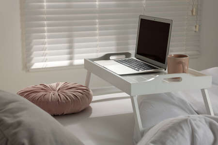 White tray table with laptop and cup of drink on bed indoors