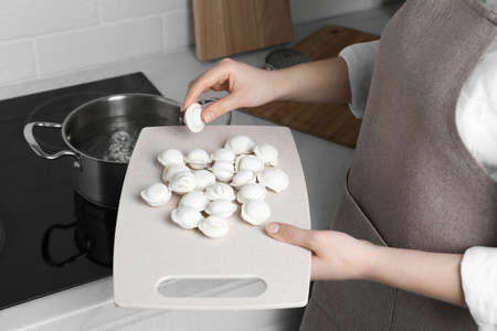 Woman putting frozen dumplings into saucepan with boiling water on cooktop in kitchen, closeup