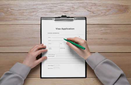 Woman filling visa application form on wooden table, top view
