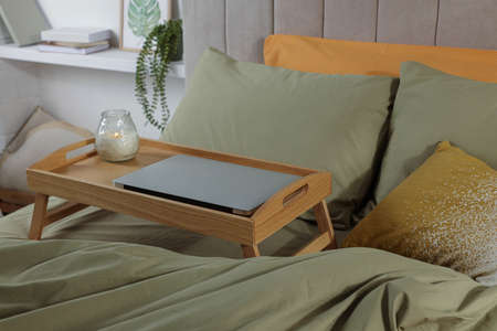 Wooden tray with modern laptop and burning candle on bed indoorsの写真素材