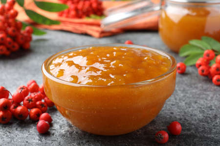 Delicious rowan jam in glass bowl and berries on gray table, closeup