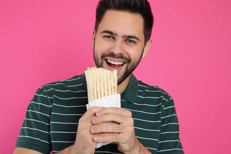 Young man eating tasty shawarma on pink background