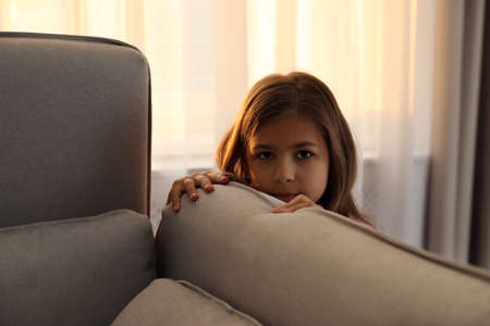Sad little girl hiding behind sofa at home. domestic violence conceptの写真素材