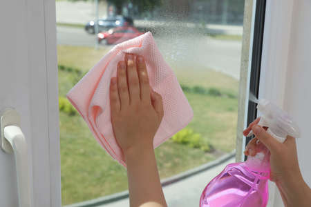 Young woman cleaning window glass with rag and detergent at home, closeup