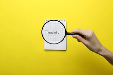 Woman holding magnifying glass over sheet of paper with word Translate on yellow background, top viewの写真素材