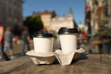 Cardboard takeaway coffee cups with plastic lids and holder on wooden table in city