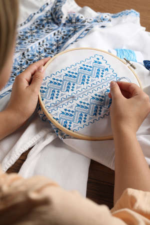 Woman embroidering white shirt with blue thread on wooden table, closeup. Ukrainian national clothes