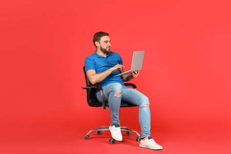 Young man with laptop sitting in comfortable office chair on red backgroundの写真素材
