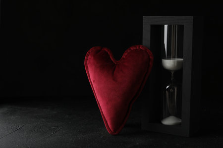 Heart shaped cushion and hourglass on black stone table, space for text. Relationship problems...