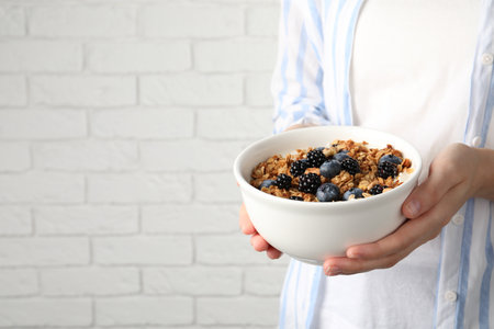 Woman holding bowl of oatmeal porridge with berries near white brick wall, closeup. Space for textの写真素材