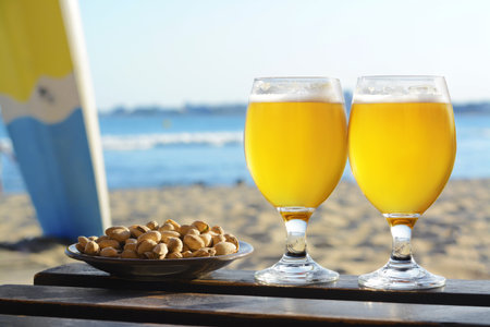 Glasses of cold beer and pistachios on wooden table near sea, space for textの写真素材