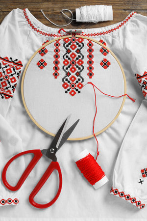 Shirt with red embroidery design in hoop, needle, scissors and threads on wooden table, flat lay. National Ukrainian clothingの写真素材