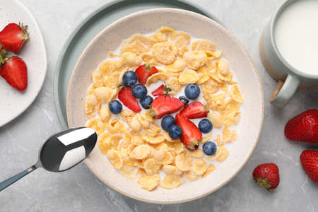Bowl of tasty crispy corn flakes with milk and berries on light gray table, flat lay