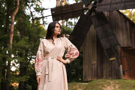 Beautiful woman wearing embroidered dress near old wooden mill in countryside. Ukrainian national clothesの写真素材