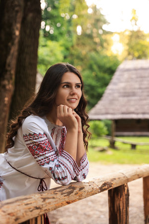 Beautiful woman wearing embroidered shirt near wooden railing in countryside. Ukrainian national clothesの写真素材