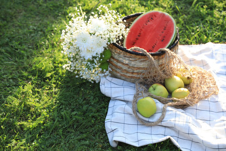 Picnic blanket with tasty fruits, beautiful flowers and basket on green grass outdoorsの写真素材