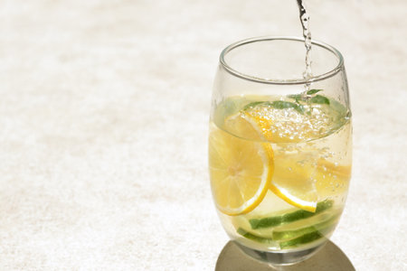 Pouring water into glass with lemon slices and mint at light gray table, closeup. Space for text