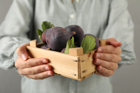Woman holding wooden crate with tasty raw figs on light gray background, closeup