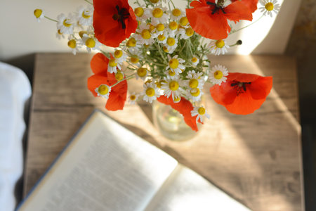 Beautiful bouquet of poppies and chamomiles near open book indoors, focus on flowersの写真素材