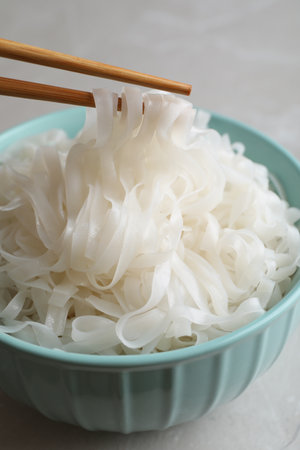 Taking rice noodles with chopsticks from bowl at light gray table, closeup