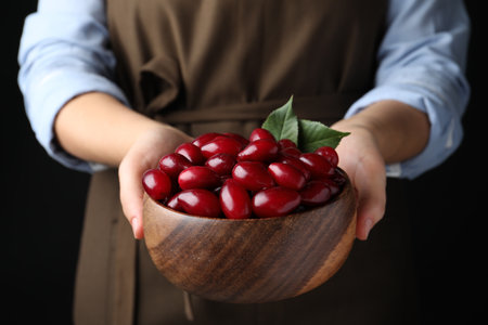 Woman with wooden bowl of fresh ripe dogwood berries on black background, closeupの写真素材