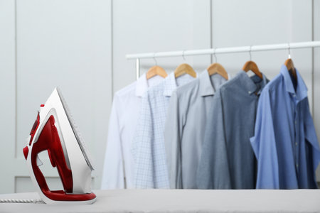 Modern iron on light table near rack with clean shirts indoors. Space for textの写真素材