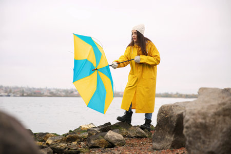 Woman in yellow raincoat with umbrella caught in gust of wind near river