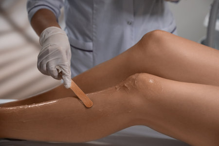 Professional cosmetologist applying gel on client's leg before laser epilation procedure in salon,...