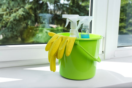 Green bucket with gloves and spray bottles of detergents on window sill indoors, space for text. cleaning suppliesの写真素材