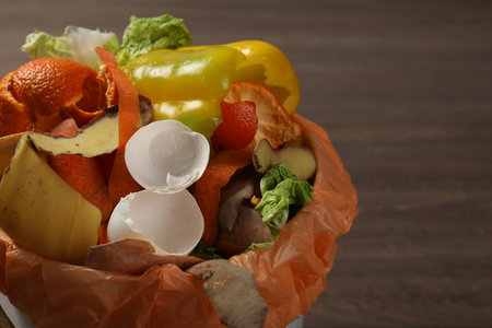Trash bin with organic waste for composting on wooden background, closeup