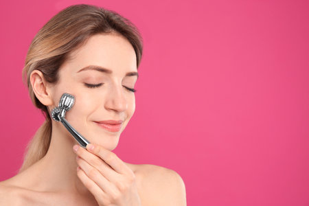 Young woman using metal face roller on pink background, space for textの写真素材