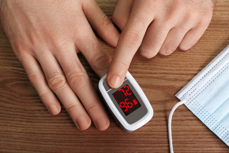 Man using pulse oximeter for oxygen level testing at wooden table with medical mask, top view