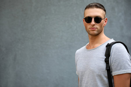 Handsome young man in stylish sunglasses with backpack near gray wall, space for text