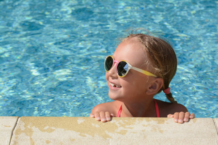 Happy cute little girl with sunglasses at the edge of swimming pool on sunny day. Space for textの写真素材