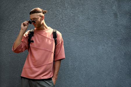 Handsome young man in stylish sunglasses and backpack near gray wall, space for text