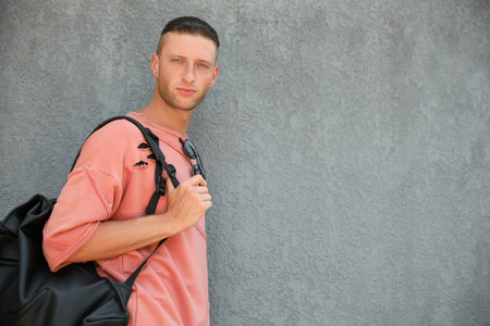 Handsome young man with stylish sunglasses and bag near gray wall, space for text