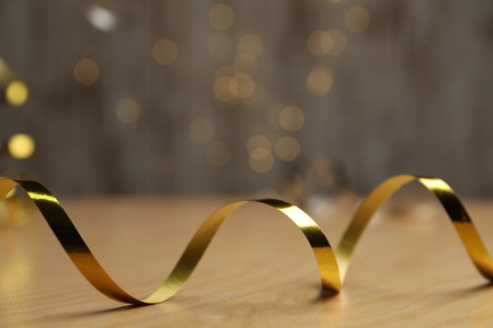 Shiny golden serpentine streamer on wooden table against blurred lights, closeup. Space for text