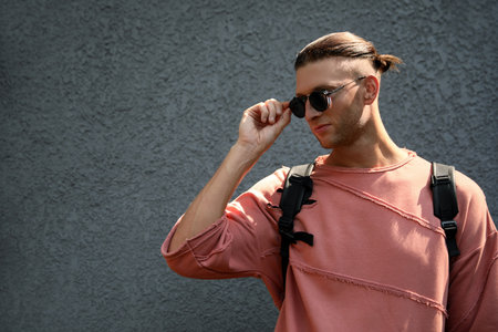 Handsome young man in stylish sunglasses and backpack near gray wall, space for text