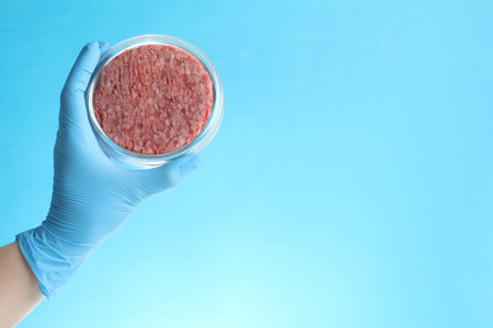 Scientist holding Petri dish with minced lab grown meat on light blue background, closeup. Space for textの写真素材