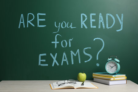 Different stationery and alarm clock on wooden table near chalkboard with phrase Are You Ready For Examsの写真素材