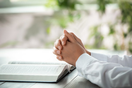 Woman holding hands clasped while praying at gray wooden table with Bible, closeup
