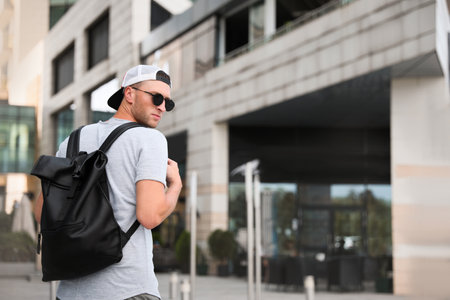 Handsome young man with stylish sunglasses and backpack near building outdoors, space for textの写真素材