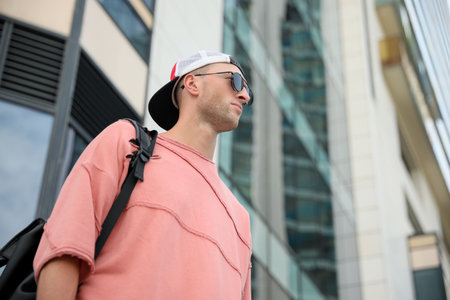 Handsome young man with stylish sunglasses and backpack near building outdoors, low angle view....